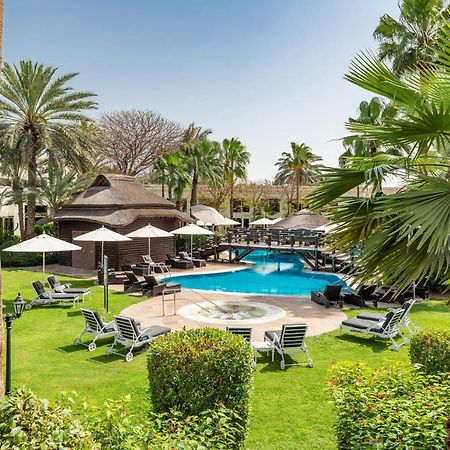 เลอ เมริเดียน ดูไบ โฮเต็ล แอนด์ คอนเฟอเรนซ์ เซ็นเตอร์ Hotel ภายนอก รูปภาพ The photo shows a serene outdoor scene featuring a swimming pool surrounded by lush greenery. There are several palm trees and shrubs providing a tropical atmosphere. Lounge chairs are arranged around the pool, along with shaded areas created by umbr