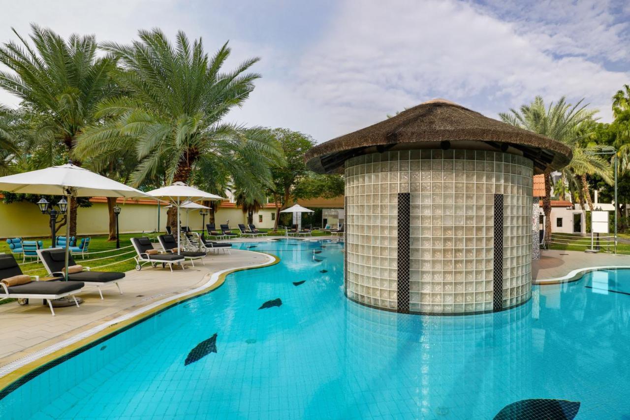 เลอ เมริเดียน ดูไบ โฮเต็ล แอนด์ คอนเฟอเรนซ์ เซ็นเตอร์ Hotel ภายนอก รูปภาพ The photo shows a luxurious pool area surrounded by palm trees. The pool has a unique design with a circular, glass-brick structure in the center, which may serve as a bar or lounge. There are lounge chairs placed around the pool, and several white u