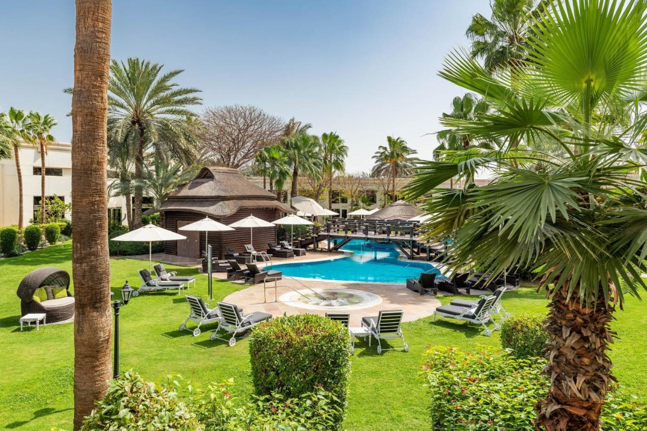 เลอ เมริเดียน ดูไบ โฮเต็ล แอนด์ คอนเฟอเรนซ์ เซ็นเตอร์ Hotel ภายนอก รูปภาพ The photo shows a serene outdoor scene featuring a swimming pool surrounded by lush greenery. There are several palm trees and shrubs providing a tropical atmosphere. Lounge chairs are arranged around the pool, along with shaded areas created by umbr