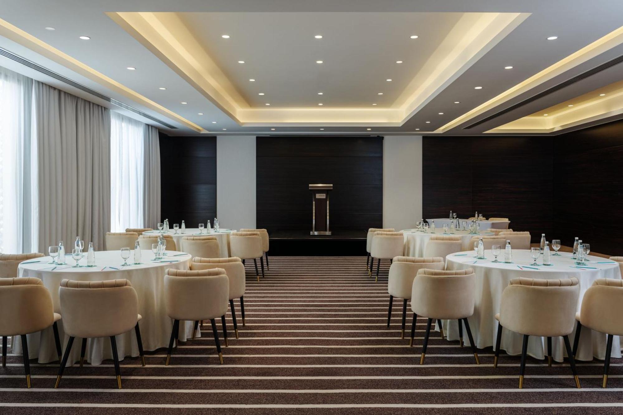 เลอ เมริเดียน ดูไบ โฮเต็ล แอนด์ คอนเฟอเรนซ์ เซ็นเตอร์ Hotel ภายนอก รูปภาพ The photo shows an elegantly designed conference or banquet room. There are round tables covered with white tablecloths, each accompanied by plush, light-colored chairs. The flooring features a striped pattern. The room is well-lit with recessed ceil