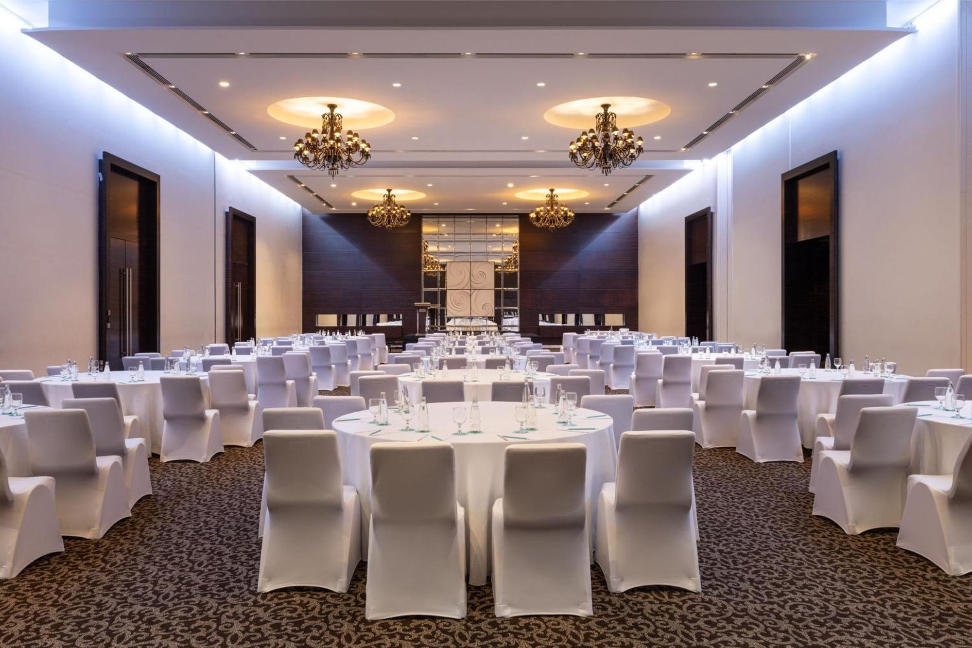 เลอ เมริเดียน ดูไบ โฮเต็ล แอนด์ คอนเฟอเรนซ์ เซ็นเตอร์ Hotel ภายนอก รูปภาพ The photo depicts an elegant banquet hall setup. The room features several round tables covered with white tablecloths, surrounded by white chair covers. The decor includes stylish chandeliers hanging from the ceiling, providing a warm and upscale am